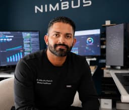 Dr. Jobby John, PharmD — founder of NimCore Women, seated in the Nimbus office in black scrubs