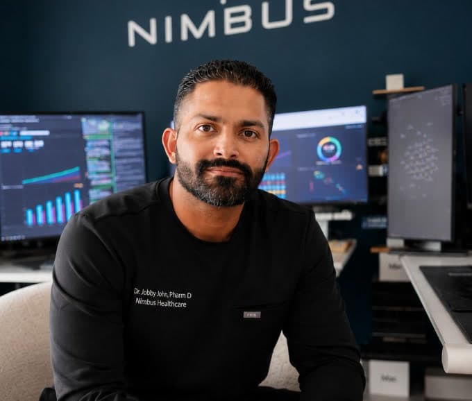 Dr. Jobby John, PharmD — founder of NimCore Women, seated in the Nimbus office in black scrubs